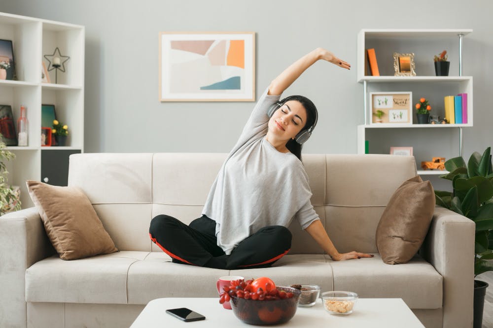 pleased young girl wearing headphones doing yoga sitting on sofa behind coffee table in living room